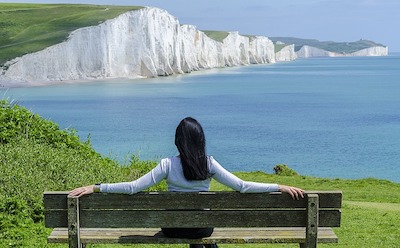 Frau sitzt entspannt mit ausgebreiteten Armen auf einer Bank und sieht auf eine weiße Küste bei hellblauen Himmel und Meerwasser